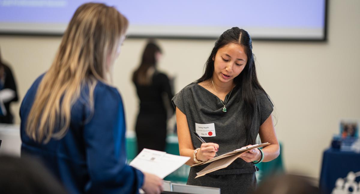 a woman with dark hair looks down at a notebook while speaking with a woman with blond hair across a table