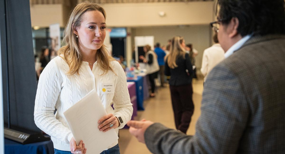 A woman with blond hair wearing a white sweater talks to a man in a suit jacket at a career fair