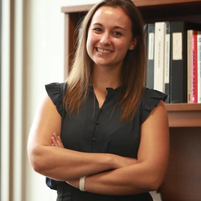 Headshot of a young woman with dark hair wearing a black sleeveless top, arms crossed, standing in front of bookshelves