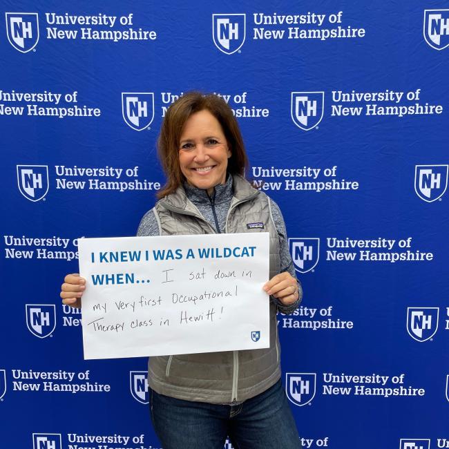 Photo of a woman smiling and holding a "I knew I was a Wildcat when..." sign in front of a UNH-branded backdrop