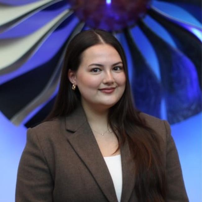 Headshot of a young woman with dark hair wearing a brown blazer, photographed against a blue and purple abstract background